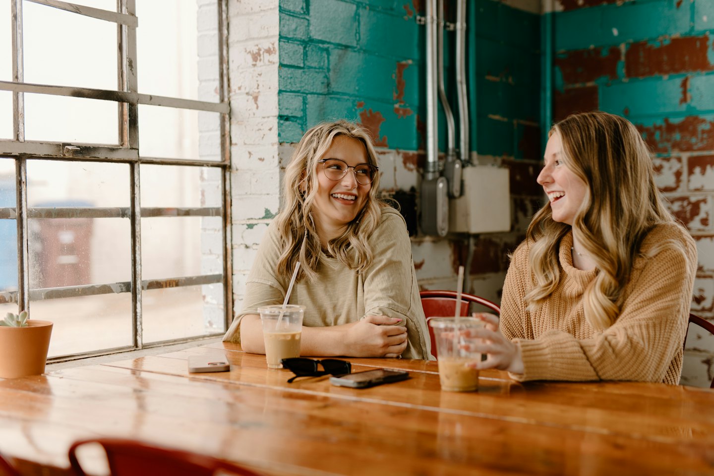 Two women laughing and drinking iced coffee at cafe table.