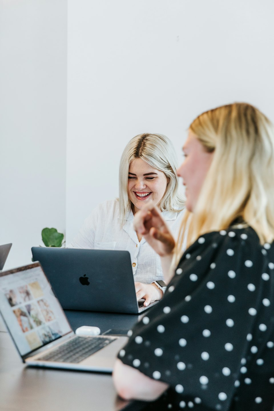 Two women collaborate and laugh while working on laptops in a bright office.